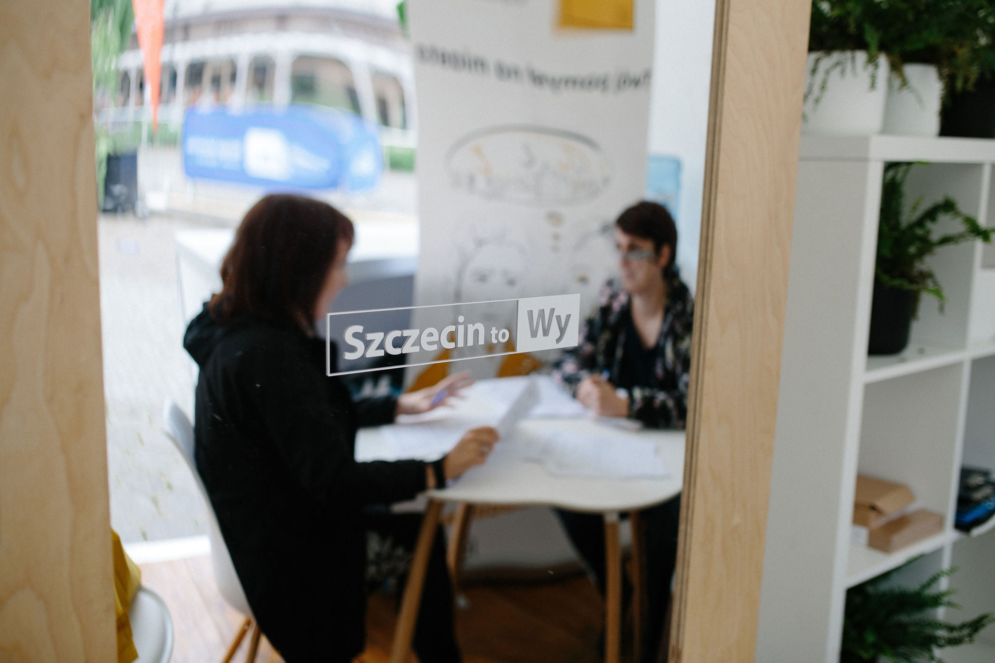 Dwie osoby siedzą przy stoliku i rozmawiają. Ich podobizny odbite są w lustrze, na którym widnieje napis: "Szczecin to Wy".